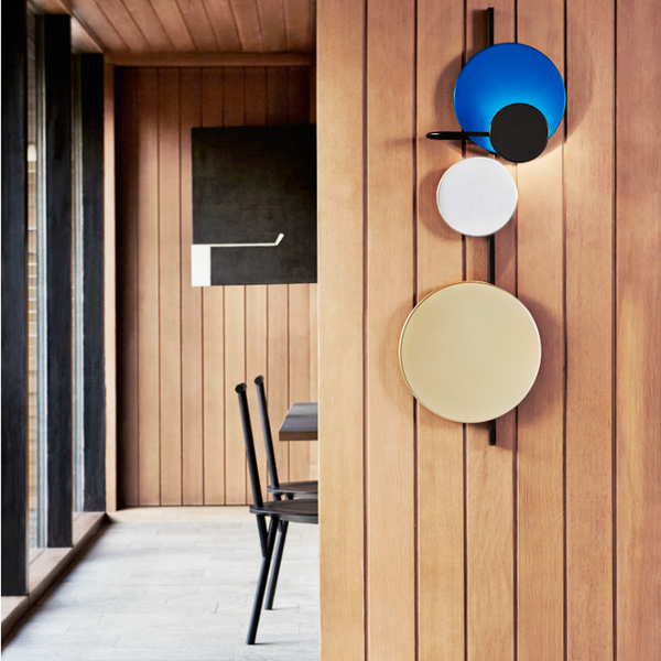 Vegglampi á panelklæddum vegg. Form lampans eru disklaga. Litir lampans eru blár, svartur, stál og gylltur.